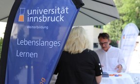 Beratungssituation im Freien, zwei Menschen stehen an einem Stehtisch. Im Vordergrund eine Beachflag der Uni Innsbruck