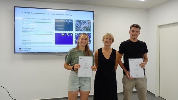 Two students holding a certificate, with Joanna in the middle, stand in front of a screen showing a slide from their project "DETECTRE: The impact of trees in cities on the environment and society"