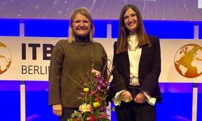 Birgit Pikkemaat und Preisträgerin Mirjam Mischi stehen auf einer Bühne, vor ihnen Blumen, im Hintergrund das Logo der ITB-Berlin