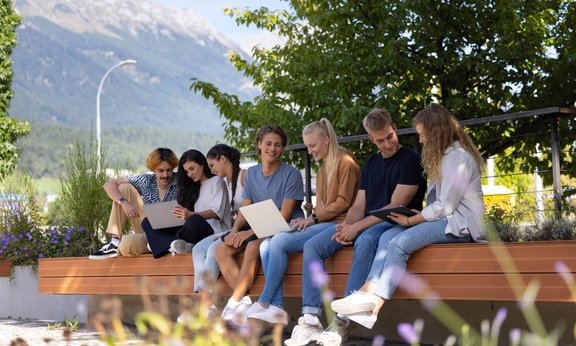 Sieben Menschen sitzen auf einer Holzbank und arbeiten in Gruppen zusammen an Laptops im Hintergrund grüne Bäume und Berge