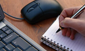 Das Bild zeigt den Ausschnitt einer Computertastatur, eine PC-Maus und eine Hand, die etwas auf einem Block notiert