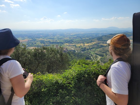 Zu sehen sind zwei Studierende von hinten. Sie blicken auf die Landschaft rund um Assisi.