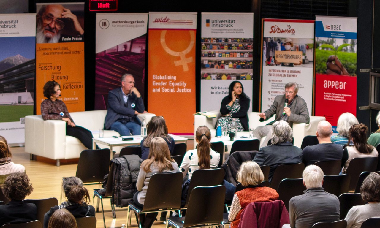 Blick in einen Veranstaltungsraum mit mehreren Personen auf einem Podium, im Vordergrund Publikum.