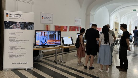 Photo of the DiSC booth which consists of a couple of tables lined up at the back of a hallway. They feature screens with videos and slides. A group of people is standing in front of the booth, talking to people from DiSC.