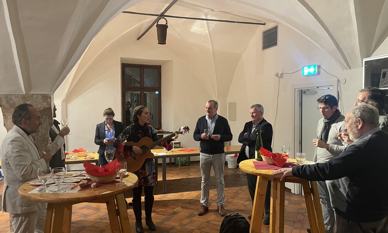 Mehrere Personen stehen in einem Raum bei einer Verkostung, in der Bildmitte spielt eine Person Gitarre.
