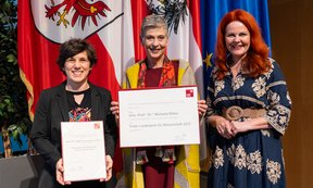 Die beiden Preisträgerinnen halten ihre Urkunden in die Kamera, rechts steht die Landesrätin, im Hintergrund die Tiroler Landesflagge.