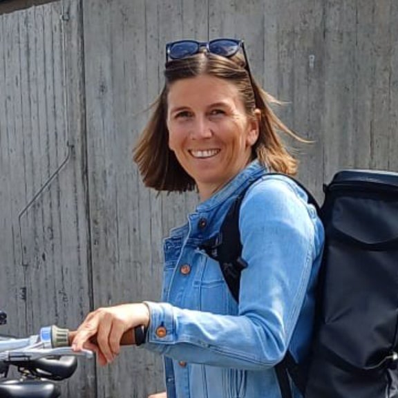 Ursula Scholl-Grissemann in Jeansjacke und mit schwarzem Rucksack stehend an einem Fahrrad