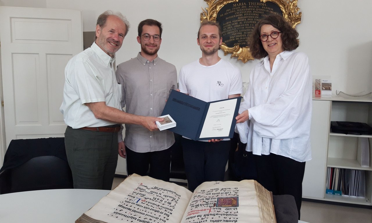 Vier Personen halten gemeinsam eine Urkunde, vor ihnen liegt ein sehr altes Buch aufgeschlagen auf einem Tisch.