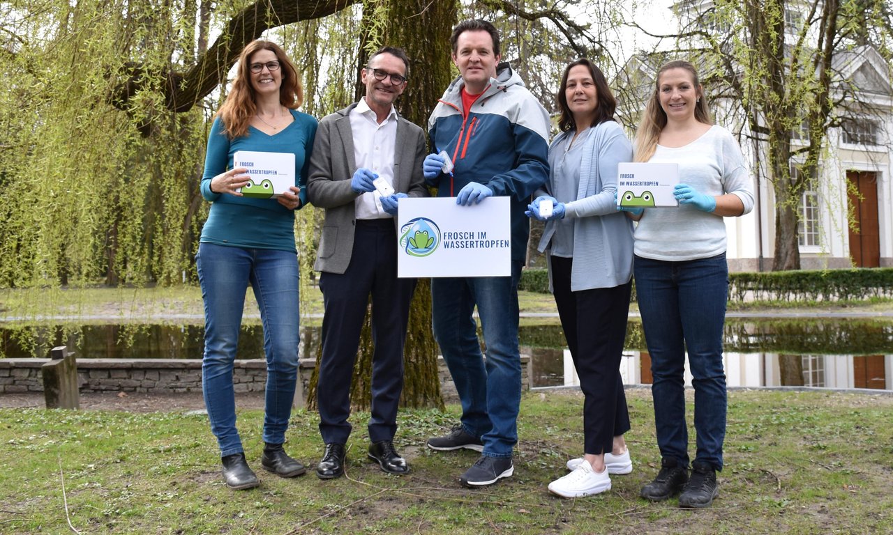 Fünf Personen stehen auf einer grünen Wiese vor einem Baum, die Person in der Mitte hält ein Schild in der Hand