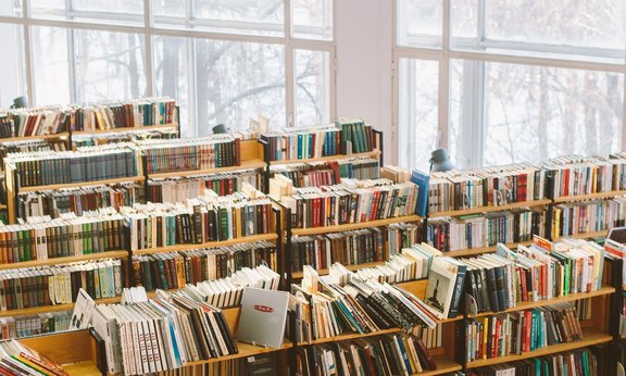 Regalreihen voller Bücher in einer Bibliothek, zu sehen sind außerdem hohe Fenster im Hintergrund.