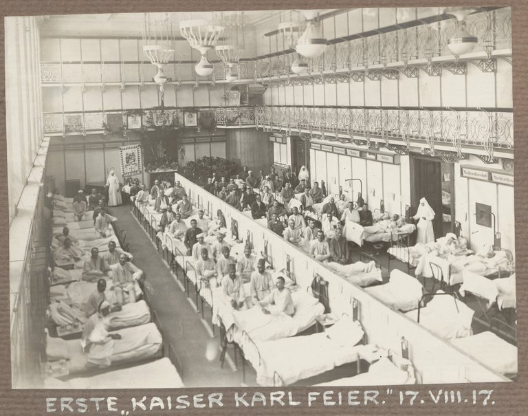 Das Foto gibt einen Blick in eine Halle, in der dicht aneinander Stahlbetten stehen, in denen Männer liegen oder sitzen, und einige Klosterschwestern vor den Türen bzw. neben einer Fahne zu sehen sind