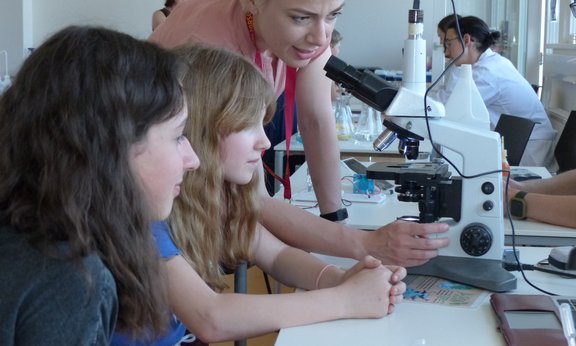 Stand bei der LNF in Mondsee: Zwei Mädchen stehen bei einem Mikroskop, einen Frau erklärt es ihnen