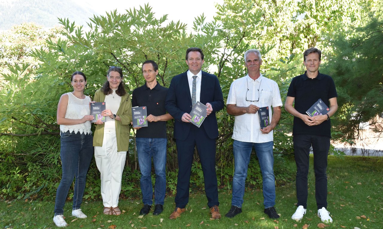 zwei Frauen und vier Männer stehen auf einer Wiese vor einem Strauch und halten kleine dunkle Bücher in der Hand.