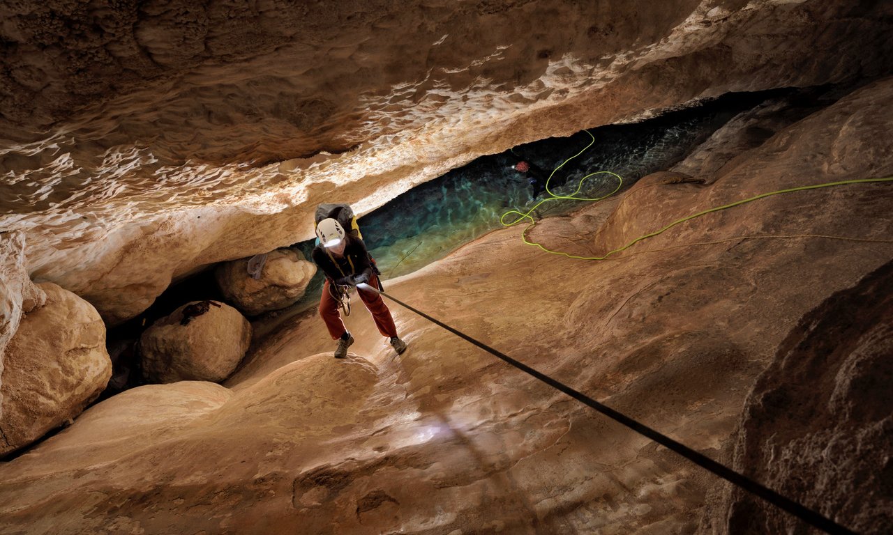 A person wearing a helmet, headlamp, and climbing harness descends a sand-colored, steep rock face in a cave using a black rope. The cave is narrow and uneven.