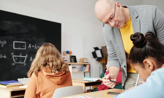 Ein Lehrer in grauem Sakko hilft einer Schülerin, seitlich ist eine weitere Schülerin zu sehen, im Hintergrund eine Tafel in einer Klasse.