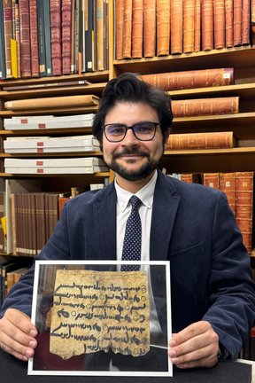 Milad Abedi an einem Schreibtisch vor einem Bücherregal sitzend, in den Händen ein durch Plexiglas geschützter beschriebener Papyrus
