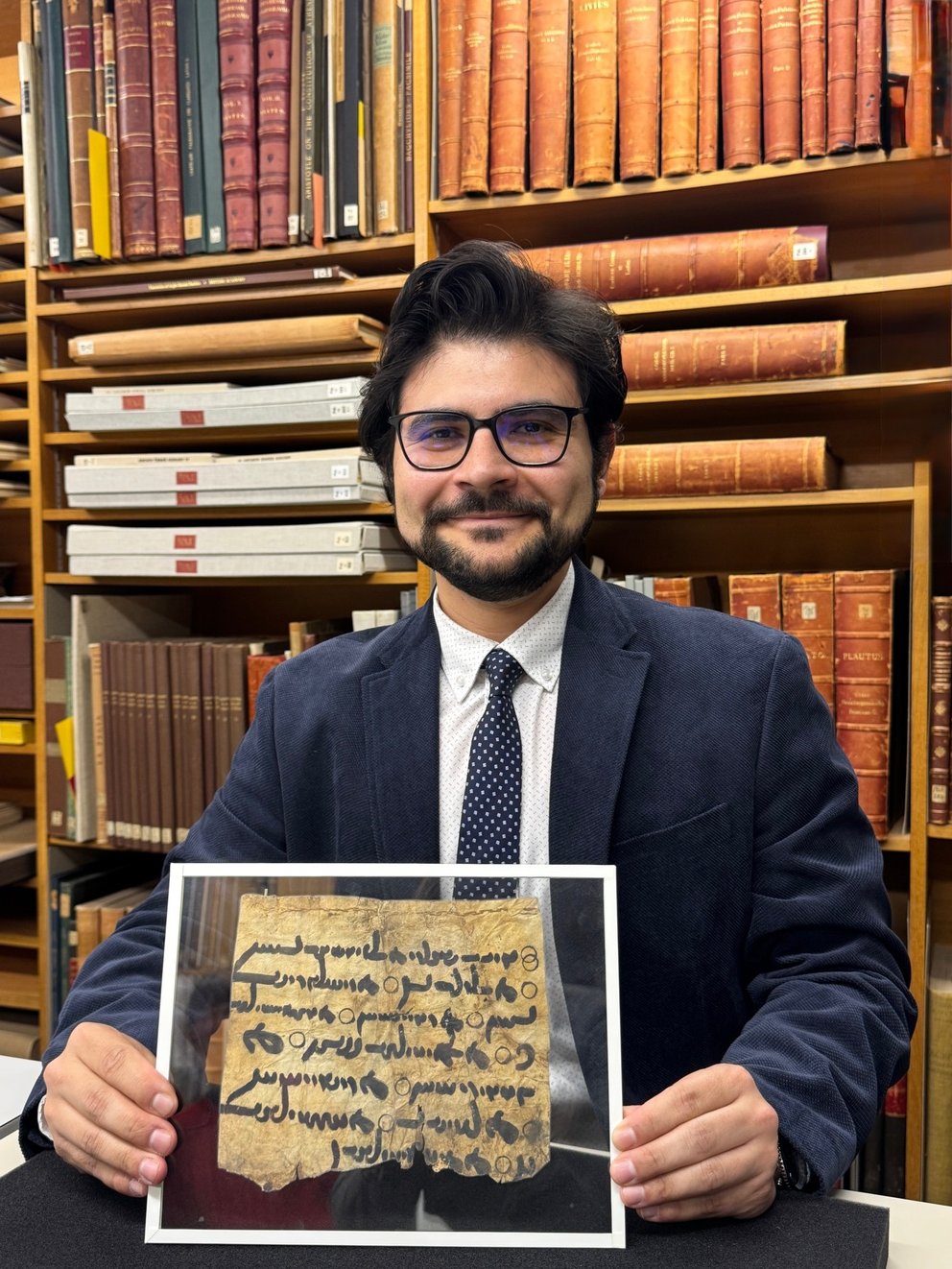 Milad Abedi an einem Schreibtisch vor einem Bücherregal sitzend, in den Händen ein durch Plexiglas geschützter beschriebener Papyrus