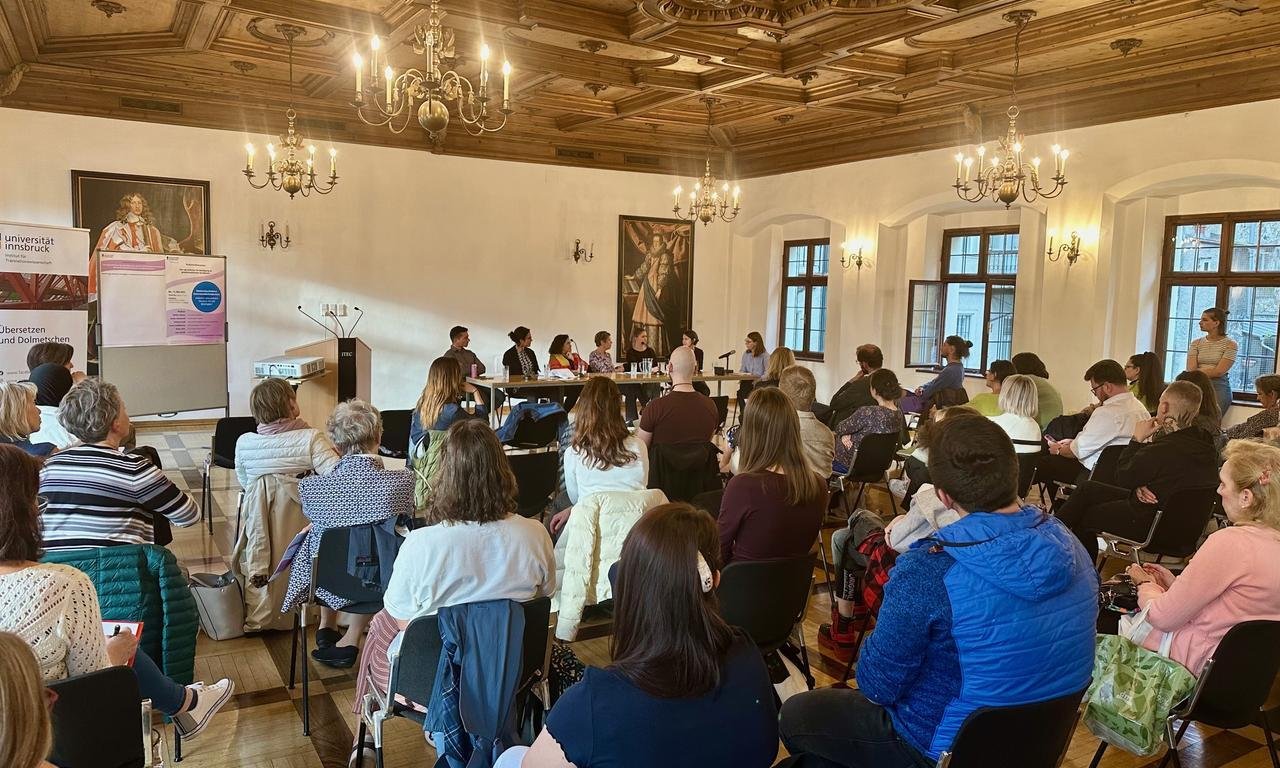 Blick von hinten in den Claudiasaal, man sieht Zuhörer:innen von hinten, Teilnehmer:innen der Podiumsdiskussion von vorne.