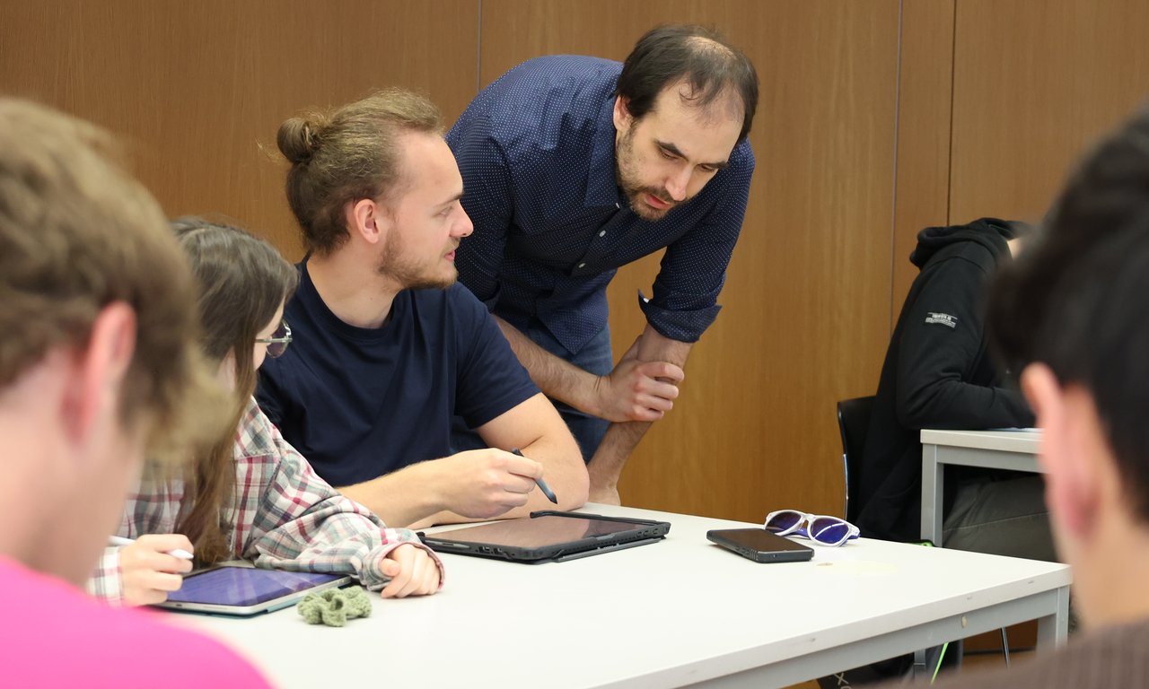 Ein Dozierender erklärt den zwei Studierenden eine Aufgabe an ihrem Tisch.