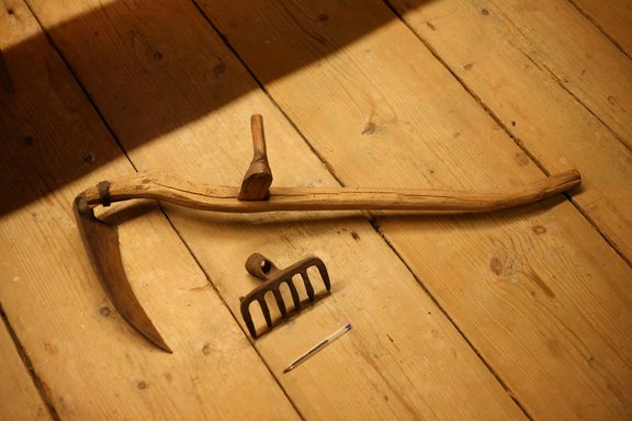 Historical short scythe about a meter long and pen wide rake head lying on a wooden parquet photographed.