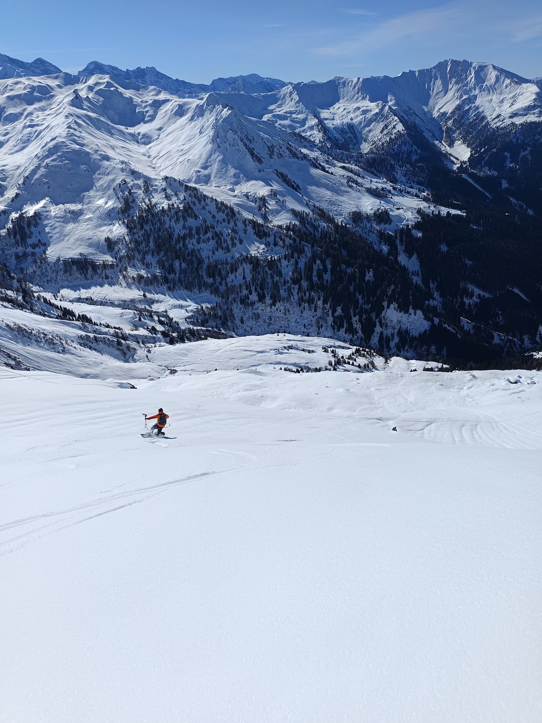 Auf dem Bild ist eine skifahrende Person zu sehen, die talwärts fährt. Hintergrund ist eine beeindruckende Kulisse schneebedeckter Berge.