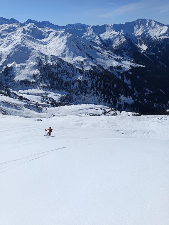 Auf dem Bild ist eine skifahrende Person zu sehen, die talwärts fährt. Hintergrund ist eine beeindruckende Kulisse schneebedeckter Berge.