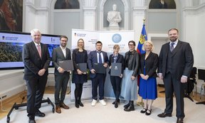 Acht Personen in formeller Kleidung stehen in einem historischen Saal vor Bannern der Universität Innsbruck und der Medizinischen Universität Innsbruck und halten Urkunden bei einer Preisverleihung.