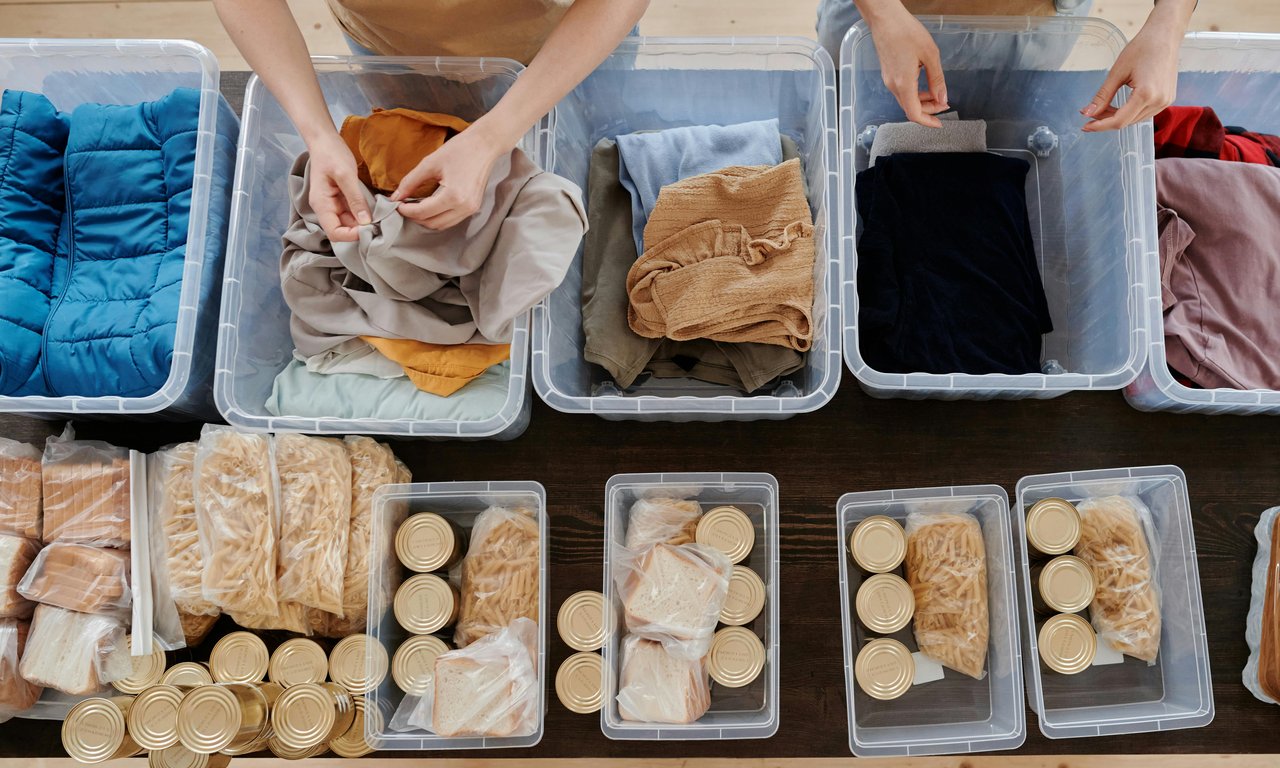 Mehrere kleine Boxen bzw. Schalen mit Spendengütern, etwa Brot oder Konserven, von oben fotografiert auf einem Tisch, dahinter die Arme von zwei Freiwilligen, die diese Boxen füllen.