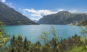 Türkisblauer Bergsee (Achensee) mit bewaldeten Hängen und hohen Bergen im Hintergrund unter klarem, sonnigem Himmel.