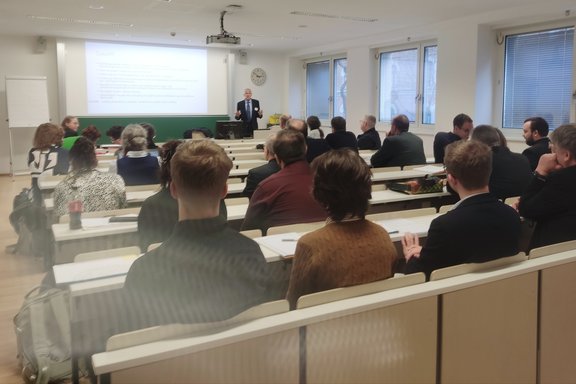 Zu sehen ist Wilhelm Rees in einem Hörsaal. Er hält einen Vortrag. Der Saal ist gut gefüllt.