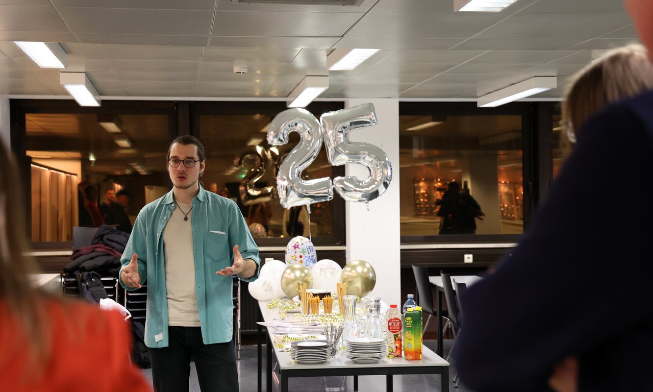 Thomas Gatt steht vor einem Tisch im Hintergrund Luftballons in der Form 25.