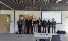 9 Personen stehen in einem Konferenzraum, im Hintergrund ist eine Powerpoint-Präsentation zu sehen