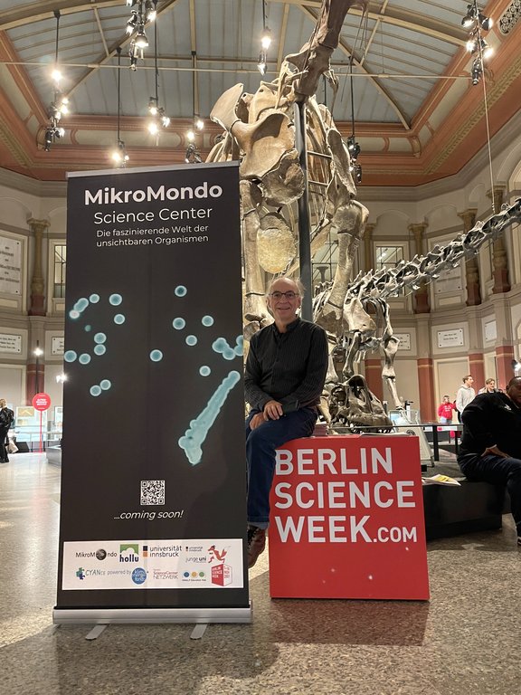 Ein Mann in einem Naturkundemuseum – hinter ihm ist ein Dinosaurier-Skelett zu sehen, außerdem ein Rollup mit dem Text „MikroMondo“, er sitzt auf einem roten, mit „Berlin Science Week“ beschrifteten Würfel.