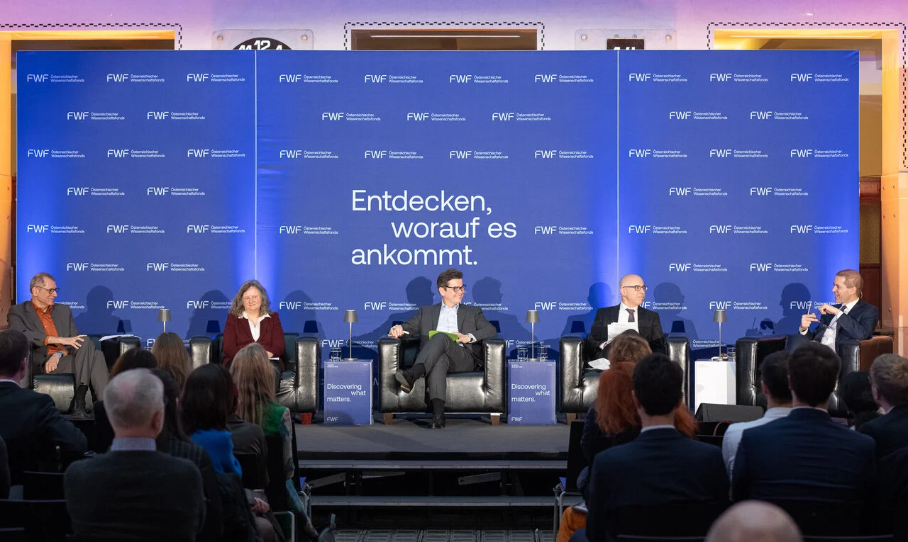 man sieht das Podium mit allen Expert:innen und im Hintergrund eine blaue Leinwand mit den FWF Schriftzeichen versehen
