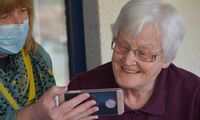 Pflegende Person zeigt älterer Dame etwas auf dem Smartphone