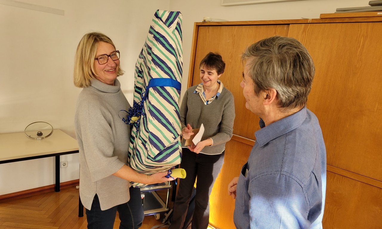 Zu sehen sind Institutsleiterin Anni Findl Ludescher mit einem großen Geschenk in den Händen zusammen mit ihren Kolleg:innen, die ihr das Geschenk überreichen.