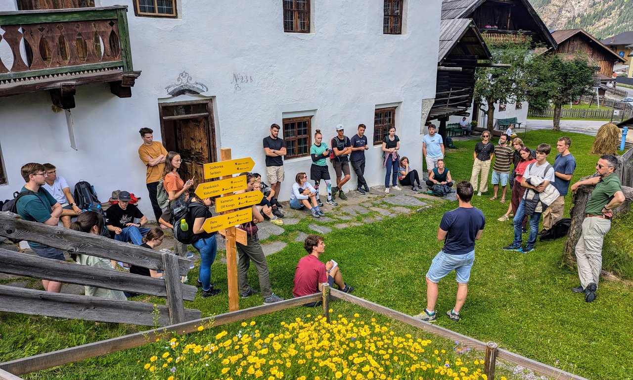 Eine Gruppe von Menschen steht verstreut auf einer Wiese vor einem alten Bauernhaus.