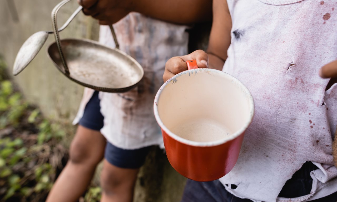 Nahaufnahme, bei denen die Hände von zwei kleinen Kindern im Mittelpunkt sind, die leere Teller halten. Das Gesicht der Kinder ist nicht zu sehen, die Kleidung macht einen ärmlichen Eindruck.