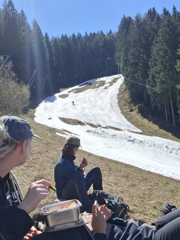 Während einer wohlverdienten Pause der Studenten im Gras fährt ein Skifahrer über die letzten Schneereste der Piste auf dem Patscherkofel