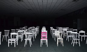 weiße Stühle vor schwarzem Hintergrund, in der Mitte ein rosa Stuhl.