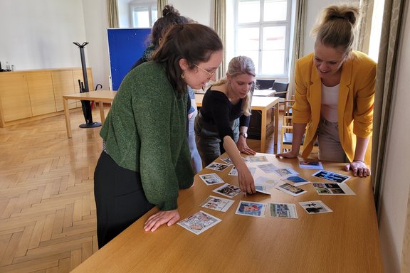 Zu sehen sind die Teilnehmer und Teilnehmerinnen des Workshops. Sie stehen an einem Tisch und analysieren Bilder.
