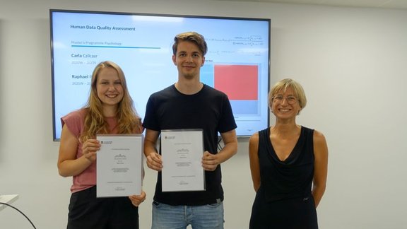 Two students holding a certificate, with Joanna on the right, stand in front of a screen showing a slide on their project "Human Data Quality Assessment".