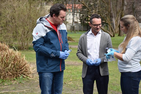 Drei Personen mit blauen Handschuhen stehen auf einer Wiesen, die Person rechts erklärt den beiden anderen etwas.