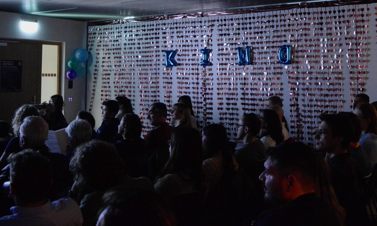Das ist Bild zeigt die Besucherinnen und Besucher des Filmabends von der Seite. Im Hintergrund ist die Wanddekoration zu erkennen: Glänzende Herzvorhänge und der Schriftzug "Kino". Das Bild ist sehr dunkel.