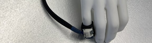 Bild zeigt ein Tool für die Kommunikation mit dem kleinen Finger. Dafür ist ein Ring mit Sensor am kleinen Finger angebracht, der über ein Kabel die Signale an eine Steuerung abgibt, die in einer Box am Unterarm festgemacht ist.