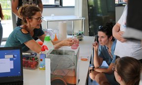 Eine Frau führt ein Experiment an einem Marmeladenglas mit roter Flüssigkeit durch. Einige Kinder und Erwachsene sehen dabei zu.