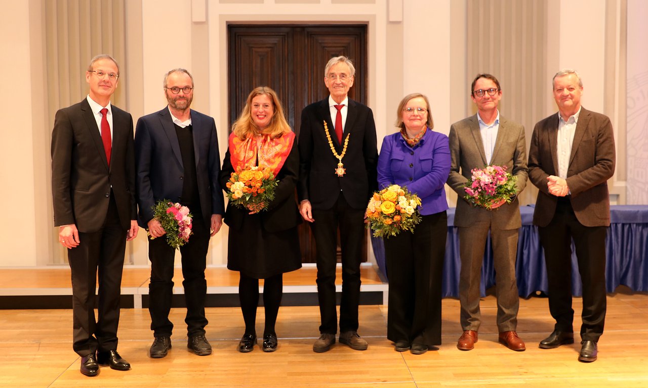 Sieben Personen stehen für ein Gruppenfoto in der Aula zusammen, einige davon haben Blumensträuße in den Händen.