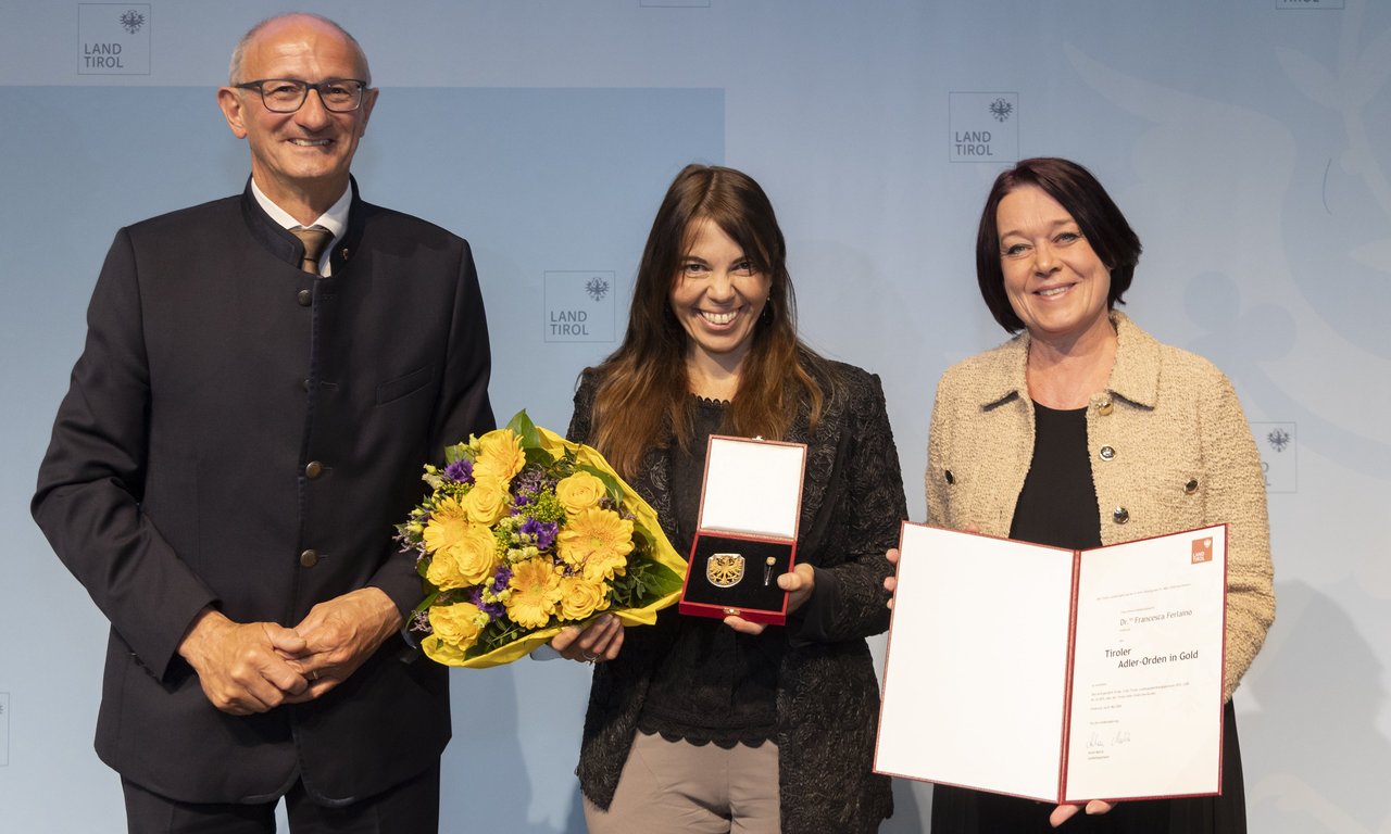 Die Preisträgerin mit Blumen und Urkunde, je eine Person links und rechts von ihr.