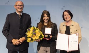Die Preisträgerin mit Blumen und Urkunde, je eine Person links und rechts von ihr.