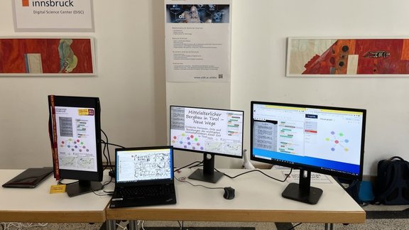 An arrangement of four screens on a table with colorful slides showing mining maps, network arrays of knowledge graphs and annotated transcriprions of texts.